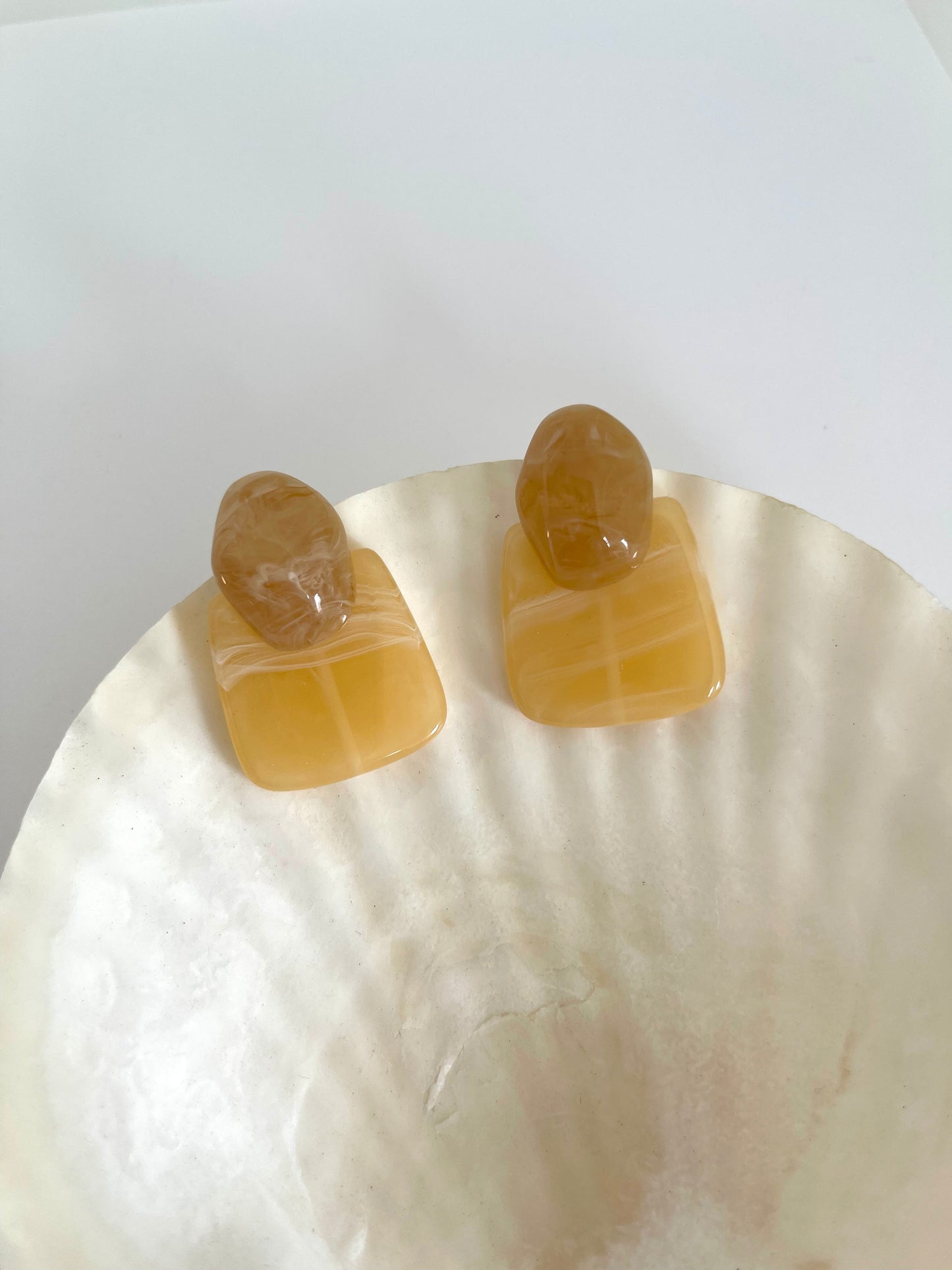 A pair of modern acrylic statement earrings in a warm amber-honey marbled design. Featuring an organic oval top and a square base, these lightweight, glossy earrings add a chic, retro-inspired touch to any outfit. Perfect for minimalist and vintage aesthetics.